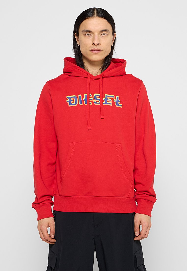 Person mit langen, dunklen Haaren, der einen roten Kapuzenpullover mit einem mehrfarbigen "DIESEL"-Logo und schwarzen Hosen trägt, steht vor einem einfarbigen Hintergrund.