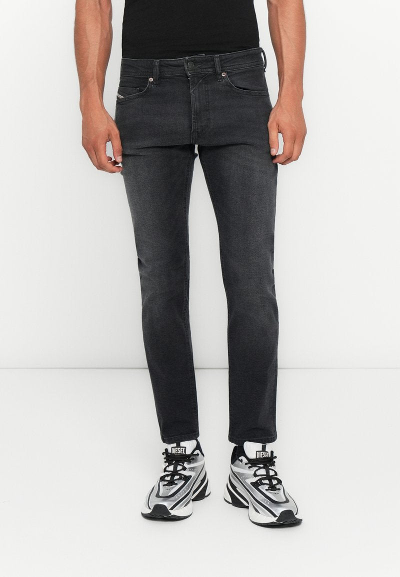 Mann in enganliegenden dunkelgrauen Jeans und silbernen, schwarzen und weißen Diesel-Sneakern, der vor einem weißen Hintergrund steht.