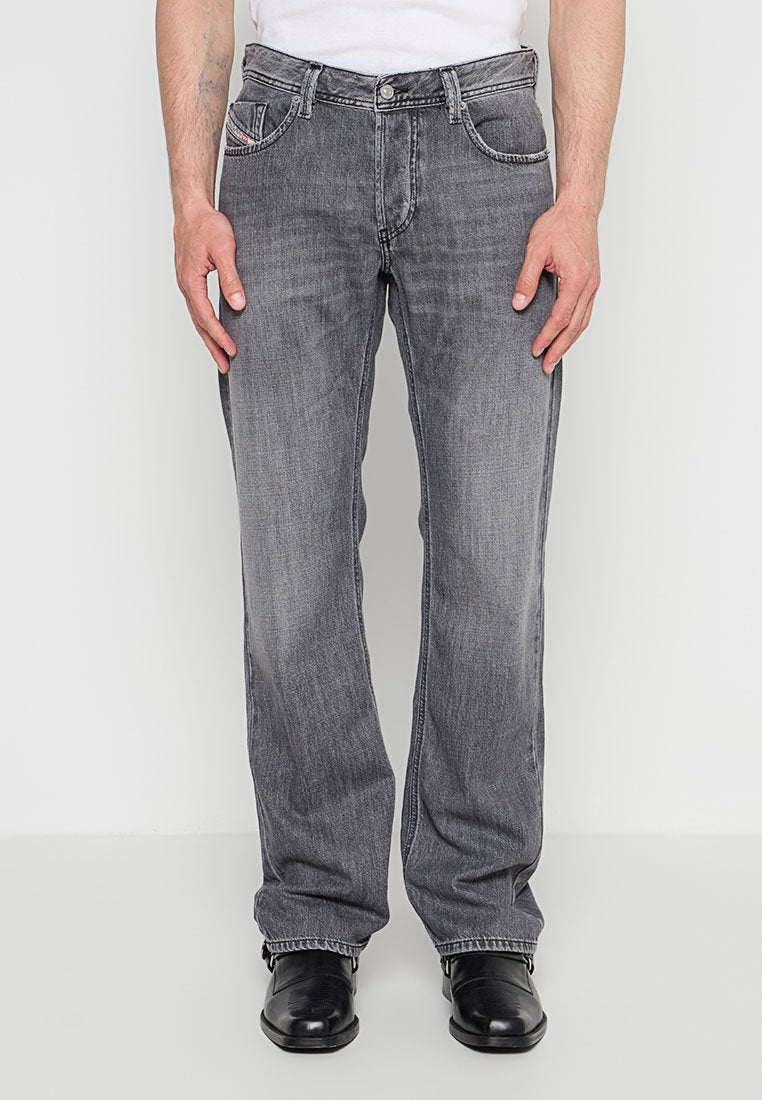 Mann trägt ausgewaschene graue Jeans mit geradem Bein, weißes Hemd eingesteckt und schwarze Lederschuhe, steht vor einem schlichten Hintergrund.