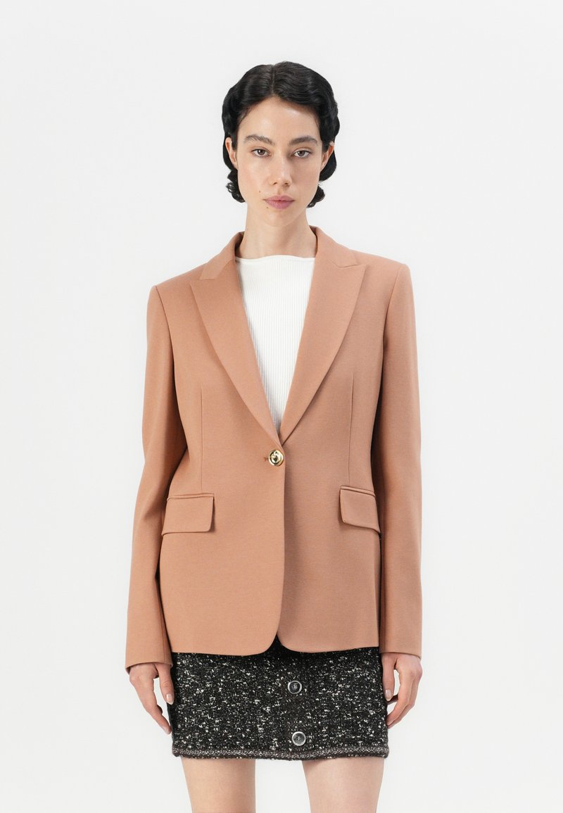 Pfirsichfarbener Blazer mit tailliertem Design, Reverskragen, zwei Fronttaschen und einem goldenen Knopf. Getragen über einem weißen Oberteil und einem schwarzen strukturierten Rock.