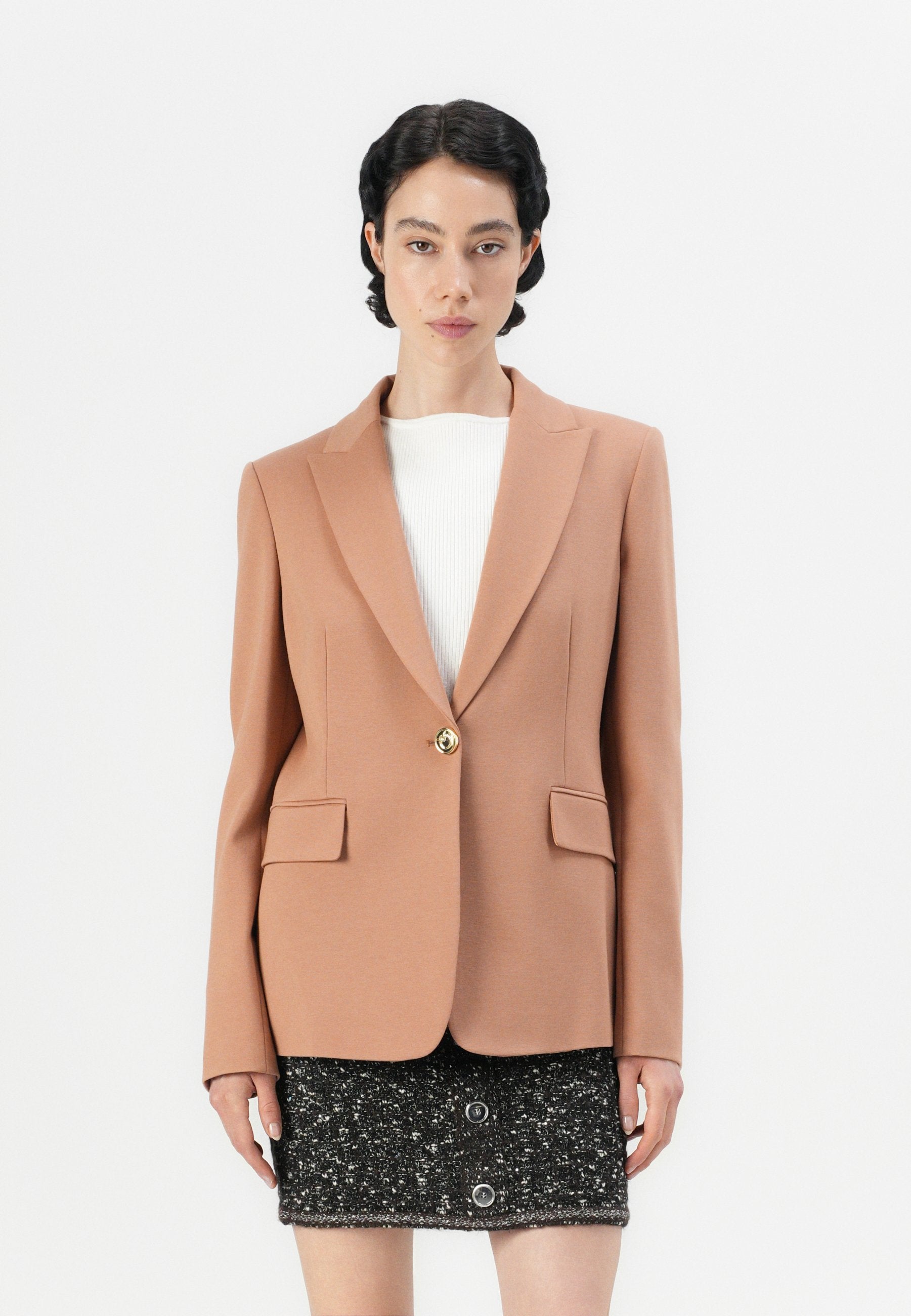 Pfirsichfarbener Blazer mit tailliertem Design, Reverskragen, zwei Fronttaschen und einem goldenen Knopf. Getragen über einem weißen Oberteil und einem schwarzen strukturierten Rock.