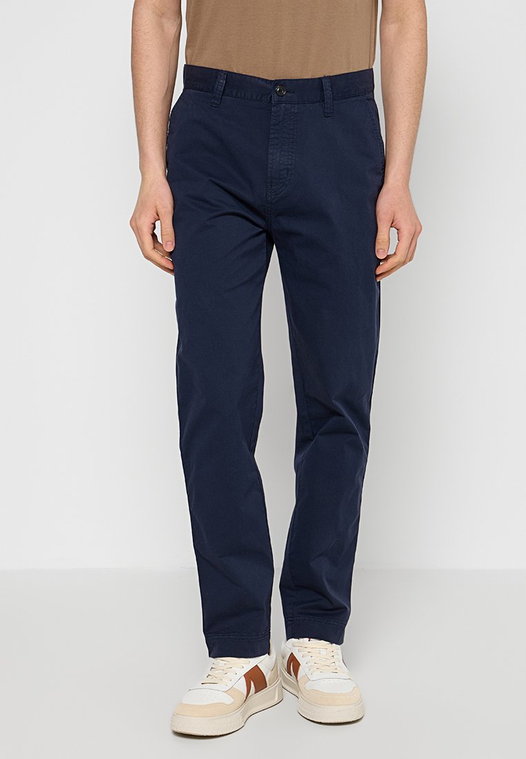Marineblaue Chinos aus Baumwolle mit schmaler Passform, ausgestattet mit Vordertaschen und einem Reißverschluss. Kombiniert mit weißen und braunen Low-Top-Sneakers.