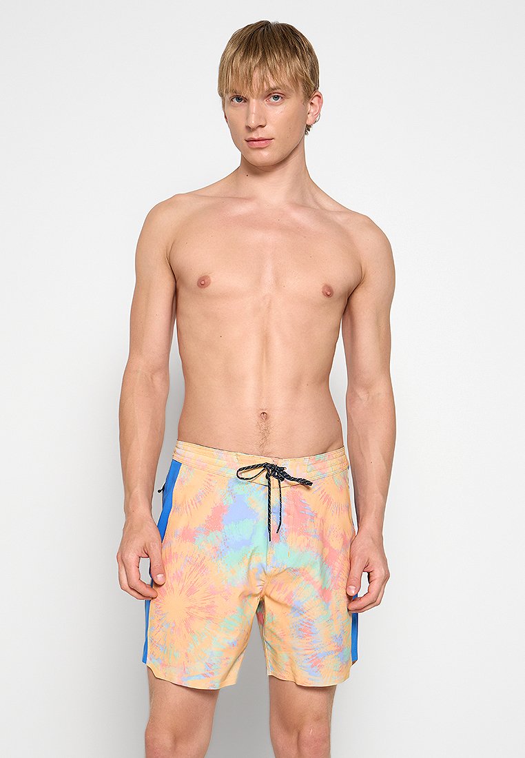 Junger Mann mit kurzem, blondem Haar, der pastellfarbene, batikfarbene Badeshorts mit blauen Seitenpanelen und einem schwarzen Kordelzug trägt.