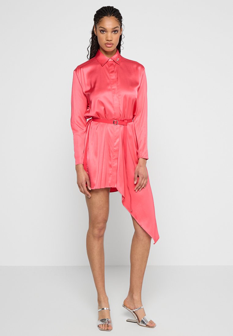 Satin rosa Hemdkleid mit Kragen, langen Ärmeln, taillierten Bund und asymmetrischem Saum. Das Modell trägt metallische Sandalen. Glatte Textur.