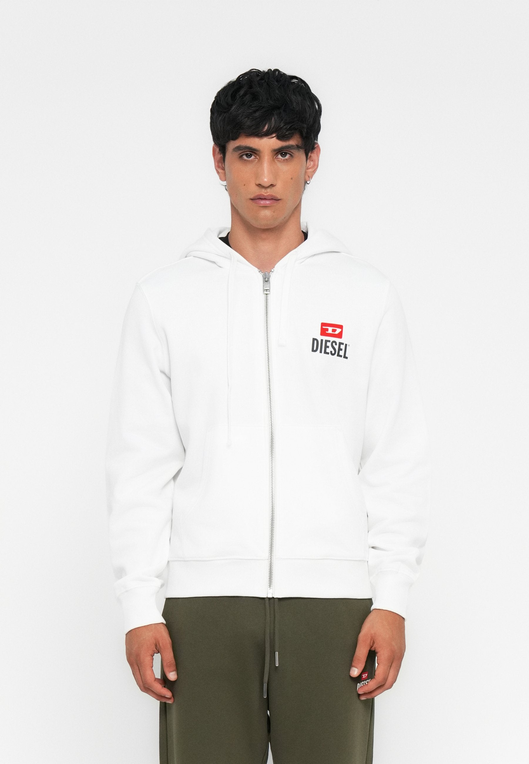 Mann in weißer Zip-Hoodie mit rotem und schwarzem Diesel-Logo und olivgrünen Hosen, steht vor einem schlichten weißen Hintergrund.