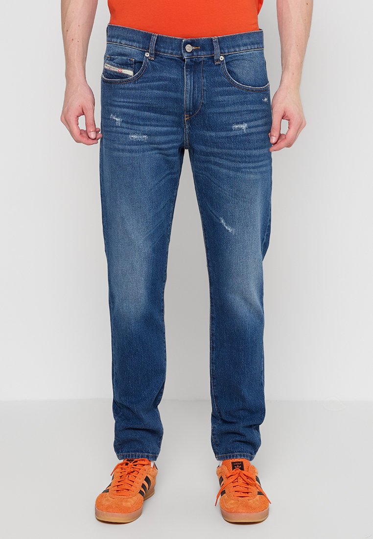 Blaue Denim-Jeans mit geradem Schnitt, die über abgenutzte Details und vier Taschen verfügen. Kombiniert mit orangefarbenen Sneakers und einem Kurzarmhemd.