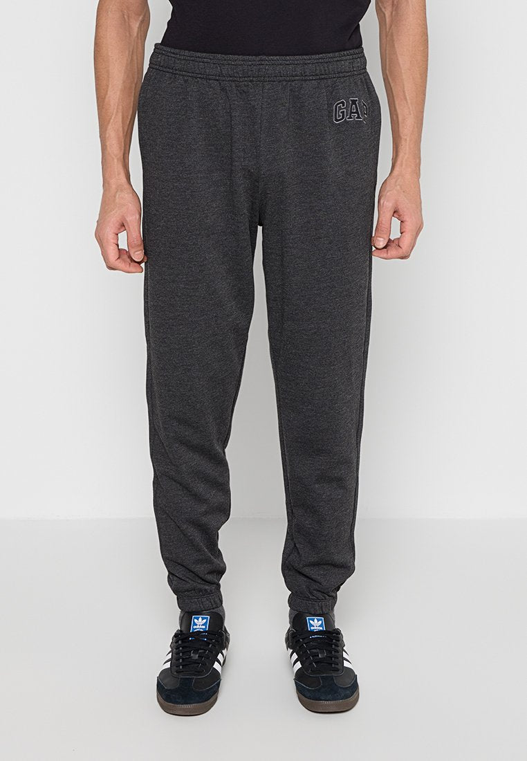 Mann trägt dunkelgraue Jogginghose mit elastischen Bündchen und GAP-Logo an der Hüfte, kombiniert mit schwarzen und weißen Adidas-Sneakern, steht auf einem schlichten Boden.