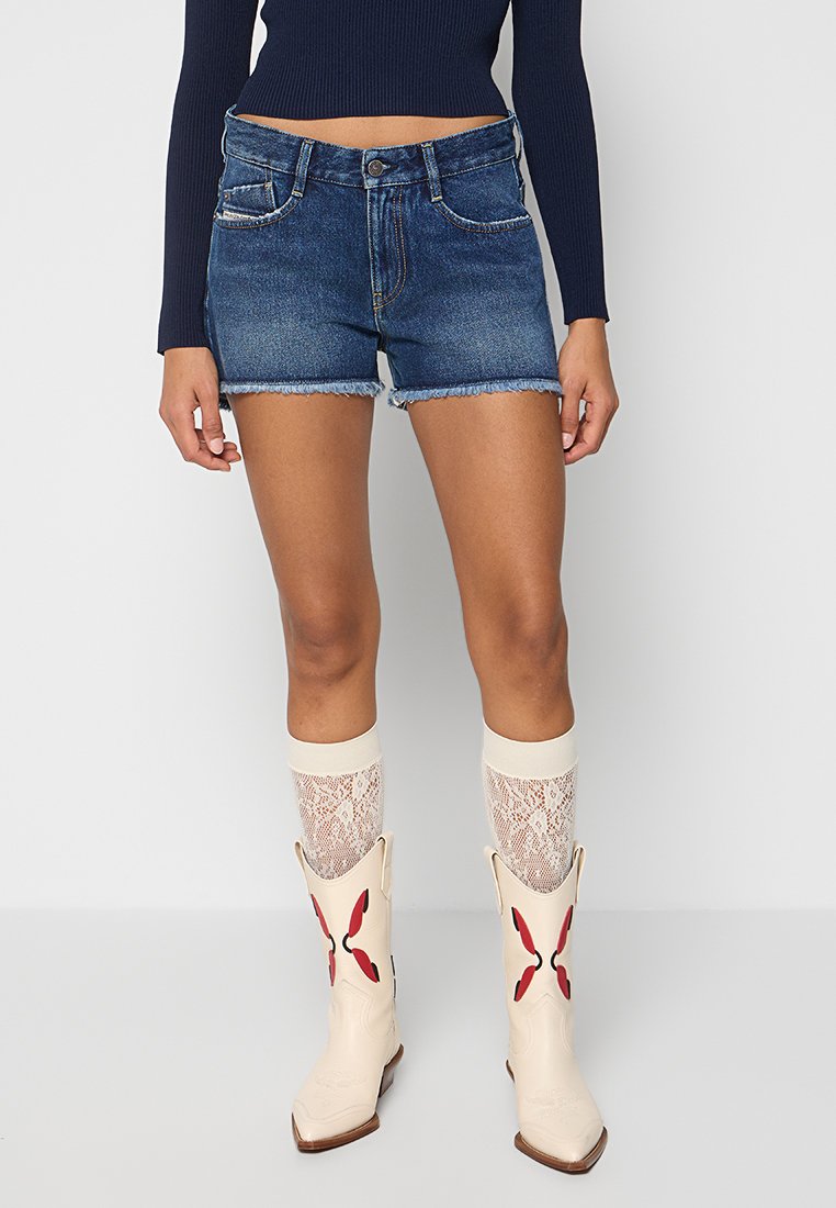 Jeans-Shorts in Mittelblau mit ausgefranstem Saum, kombiniert mit cremefarbenen Spitzenstrümpfen und cremefarbenen Cowboy-Stiefeln mit roten Akzenten.