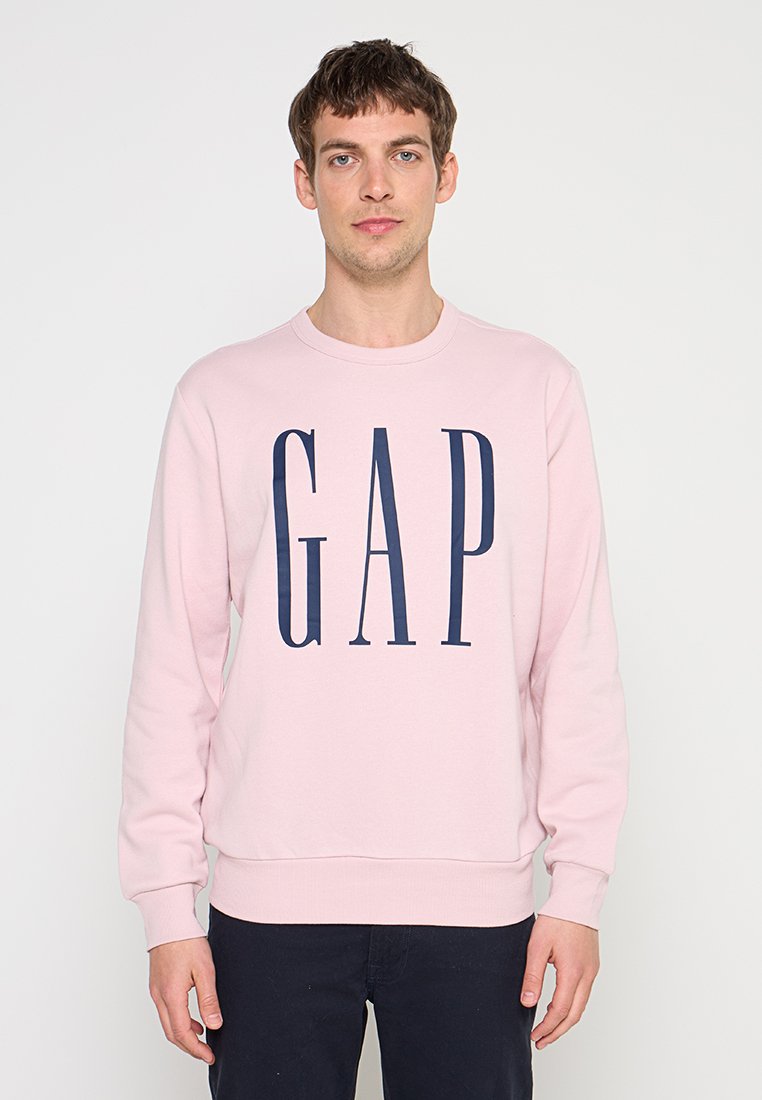 Junger Mann, der nach vorne blickt, trägt ein hellrosa Sweatshirt mit großen dunkelblauen „GAP“-Buchstaben und dunkle Hose.