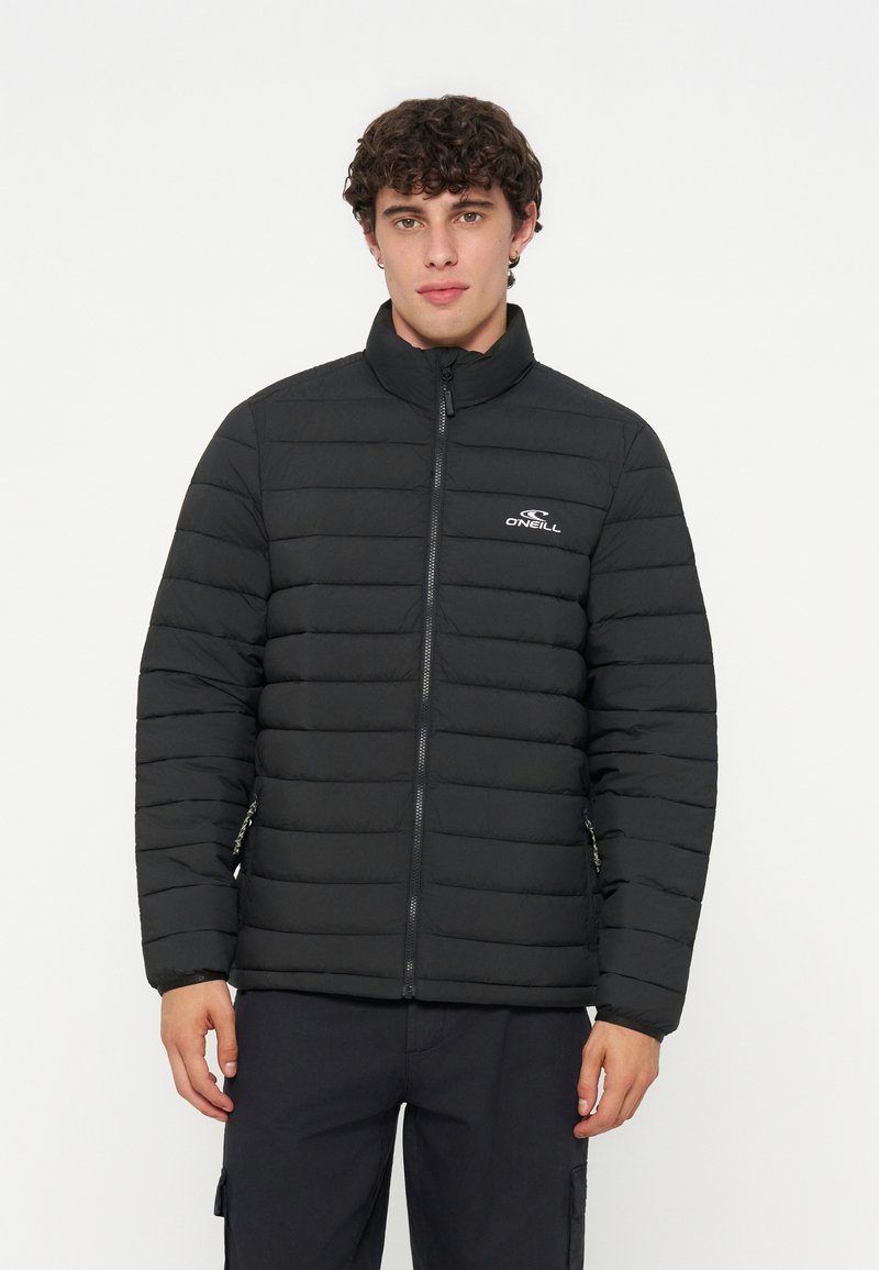 Junger Mann mit kurzen lockigen Haaren, der eine schwarze gesteppte O'Neill Jacke mit Reißverschluss und schwarze Hose trägt, vor einem schlichten weißen Hintergrund.