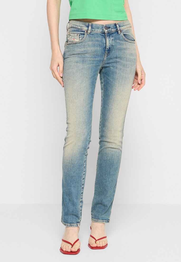 Frau in ausgewaschenen blauen Skinny-Jeans, grünem cropped Top und roten Flip-Flops, die vor einem schlichten hellen Hintergrund steht.