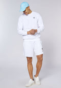 Junger Mann trägt weißen Pullover und Shorts mit blauer Kappe und weißen Turnschuhen, steht vor schlichtem hellem Hintergrund.