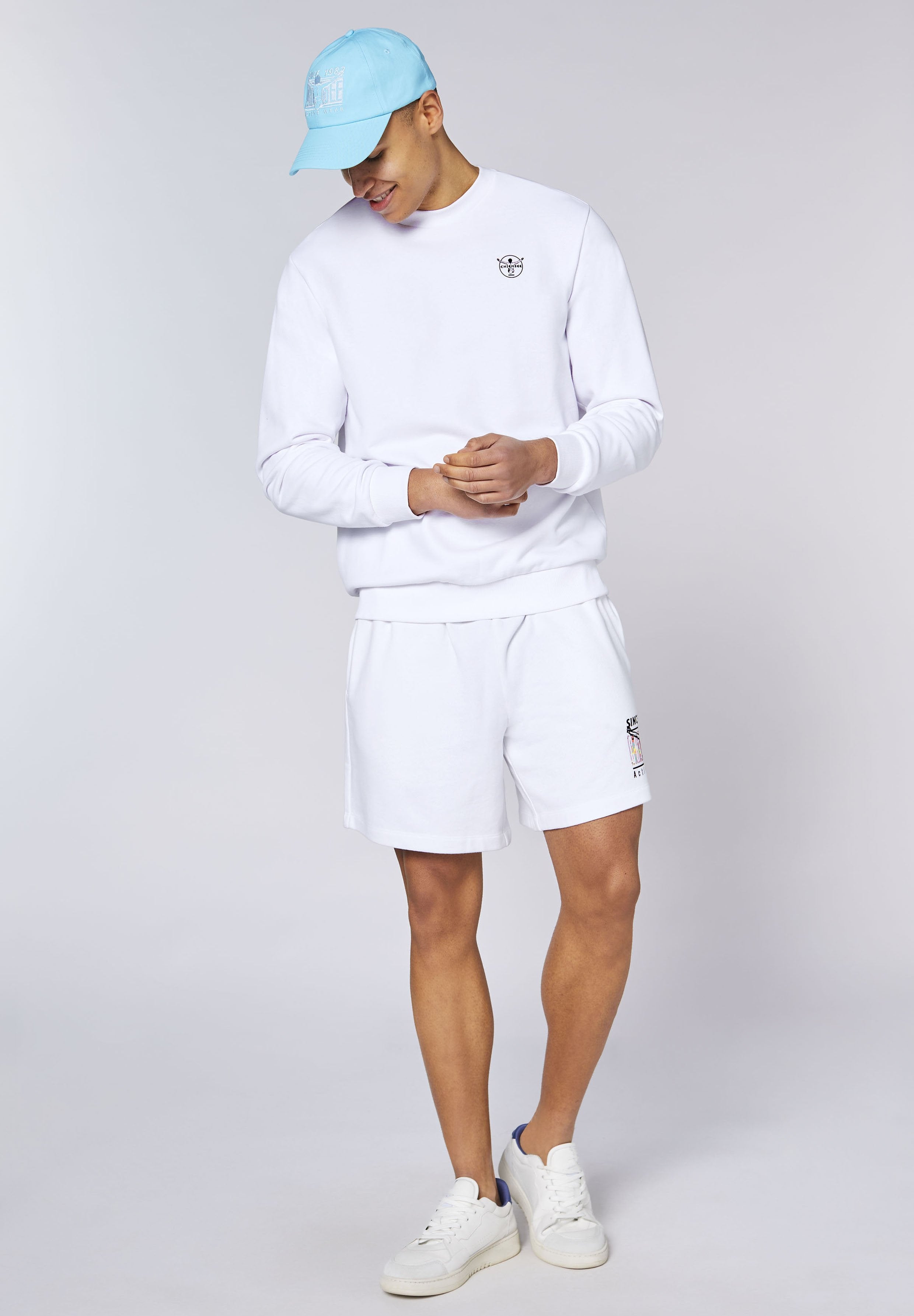 Junger Mann trägt weißen Pullover und Shorts mit blauer Kappe und weißen Turnschuhen, steht vor schlichtem hellem Hintergrund.