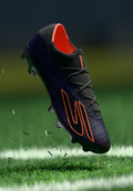 Schwarzer Fußballschuh mit orangefarbenem Innenfutter und Logo, strukturiertem Obermaterial und geformter Stollen-Sohle, in der Luft über grünem Gras aufgenommen.
