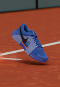 Blauer Sportschuh mit Mesh-Obermaterial, schwarzem Swoosh-Logo, strukturierter Sohle und orangefarbenen Akzenten, in der Luft über einer roten Platzoberfläche gezeigt.