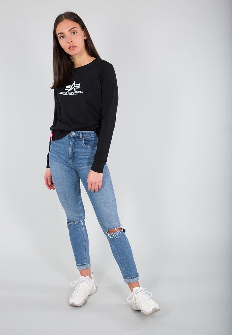 Schwarzes Langarmshirt mit "ALPHA INDUSTRIES"-Logo, kombiniert mit hellblauen, zerrissenen Jeans und weißen Sneakers, vor einem schlichten Hintergrund stehend.
