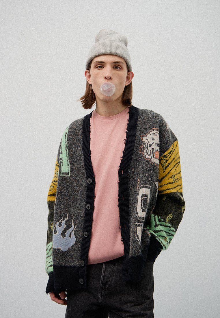 Junge Person trägt eine graue Mütze, einen gemusterten Cardigan und ein pinkes Hemd, bläst einen Kaugummi-Überzug und steht vor einem schlichten weißen Hintergrund.