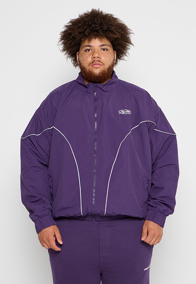 Mann mit lockigem Haar und Bart, der eine lila Zip-Jacke mit weißen Akzenten und passende lila Hosen trägt, steht vor einem einfarbigen Hintergrund.