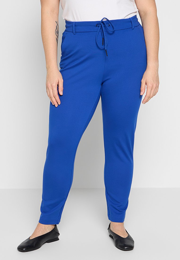 Eine Frau trägt eine leuchtend blaue, figurbetonte Hose mit einem Kordelzugbund und schwarzen flachen Schuhen, während sie vor einem schlichten hellen Hintergrund steht.