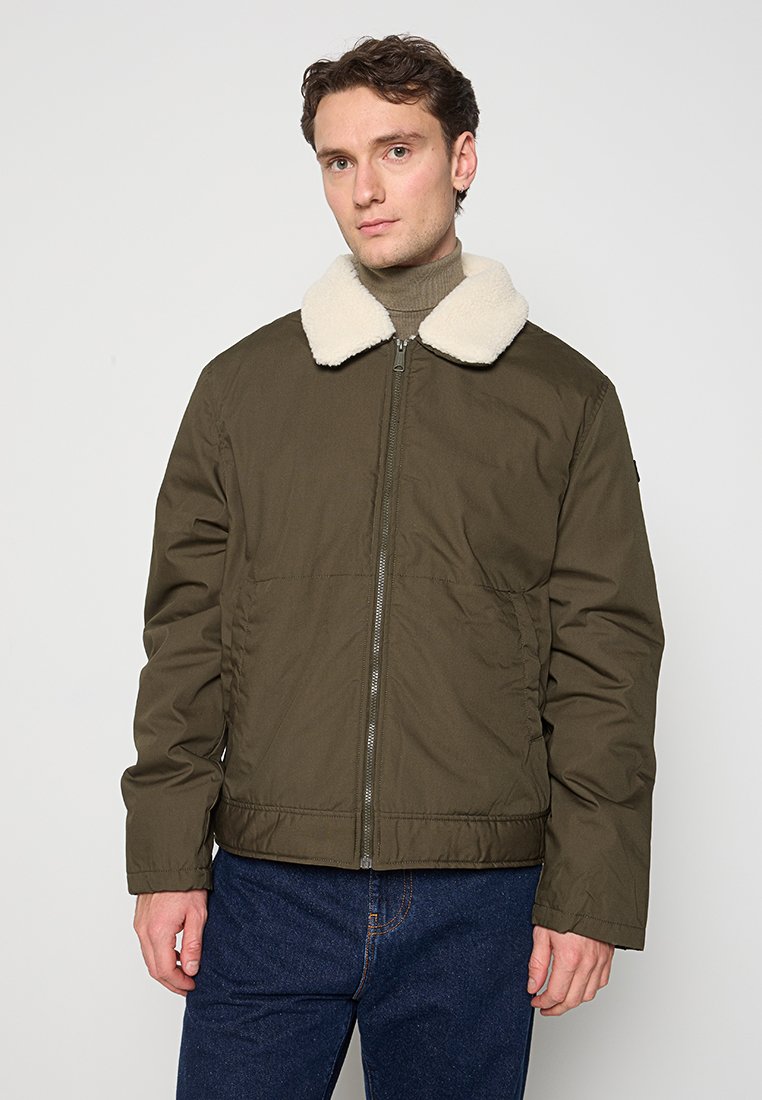 Olivgrüne Jacke mit einem weichen, weißen Kunstpelzkragen, Reißverschluss und Seitentaschen. Das Material wirkt glatt mit matter Oberfläche.