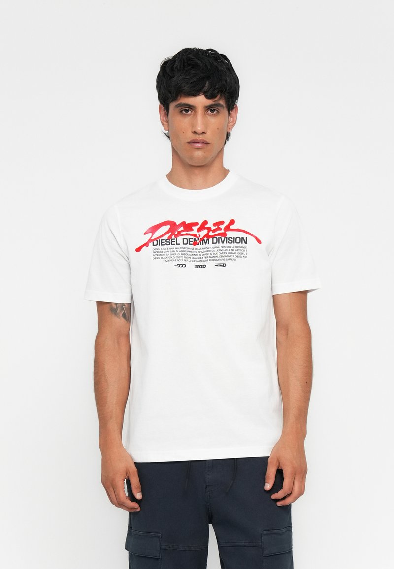 Junger Mann mit kurzen dunklen Haaren, der ein weißes Diesel Denim Division T-Shirt und dunkle Cargo-Shorts trägt, vor einem schlichten weißen Hintergrund stehend.