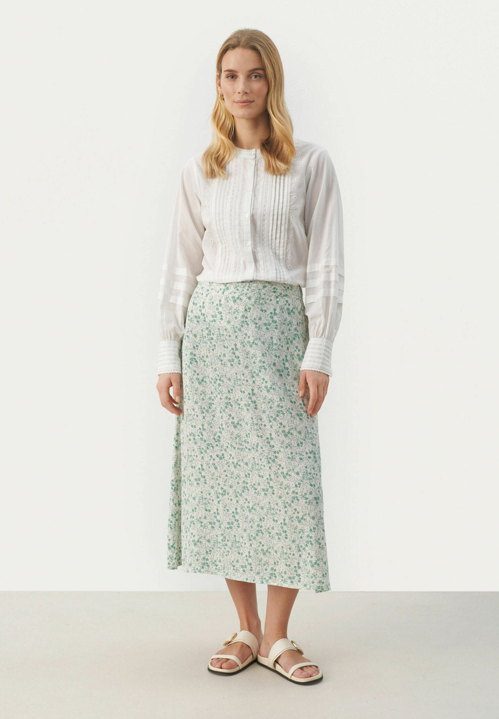 Weißes Langarmhemd mit vertikalen Falten, kombiniert mit einem grünen Maxirock mit Blumenmuster und beigen Sandalen mit goldenen Akzenten.