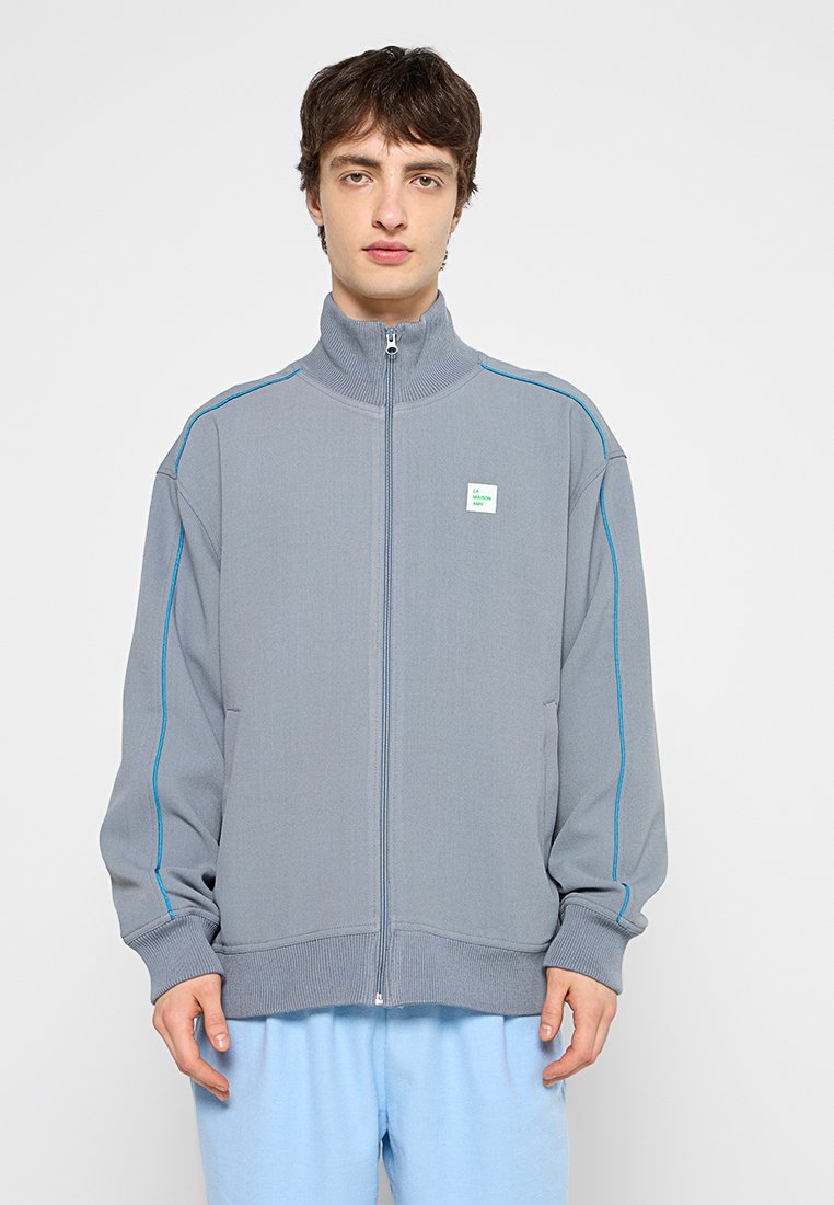 Junger Mann, der eine graue Zip-Jacke mit blauen Paspeln und eine hellblaue Hose trägt und vor einem schlichten weißen Hintergrund steht.
