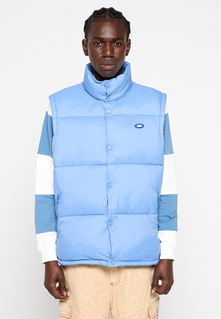 Mann mit einer hellblauen gepolsterten Weste über einem weißen und blauen Langarmhemd und beigen gemusterten Hosen, der vor einem schlichten weißen Hintergrund steht.