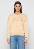 Beiger Pullover mit rundem Ausschnitt, überschnittenen Schultern und gerippten Bündchen. Auf der Vorderseite befindet sich eine metallische "MCXX"-Beschriftung. Kombiniert mit blauen Jeans.