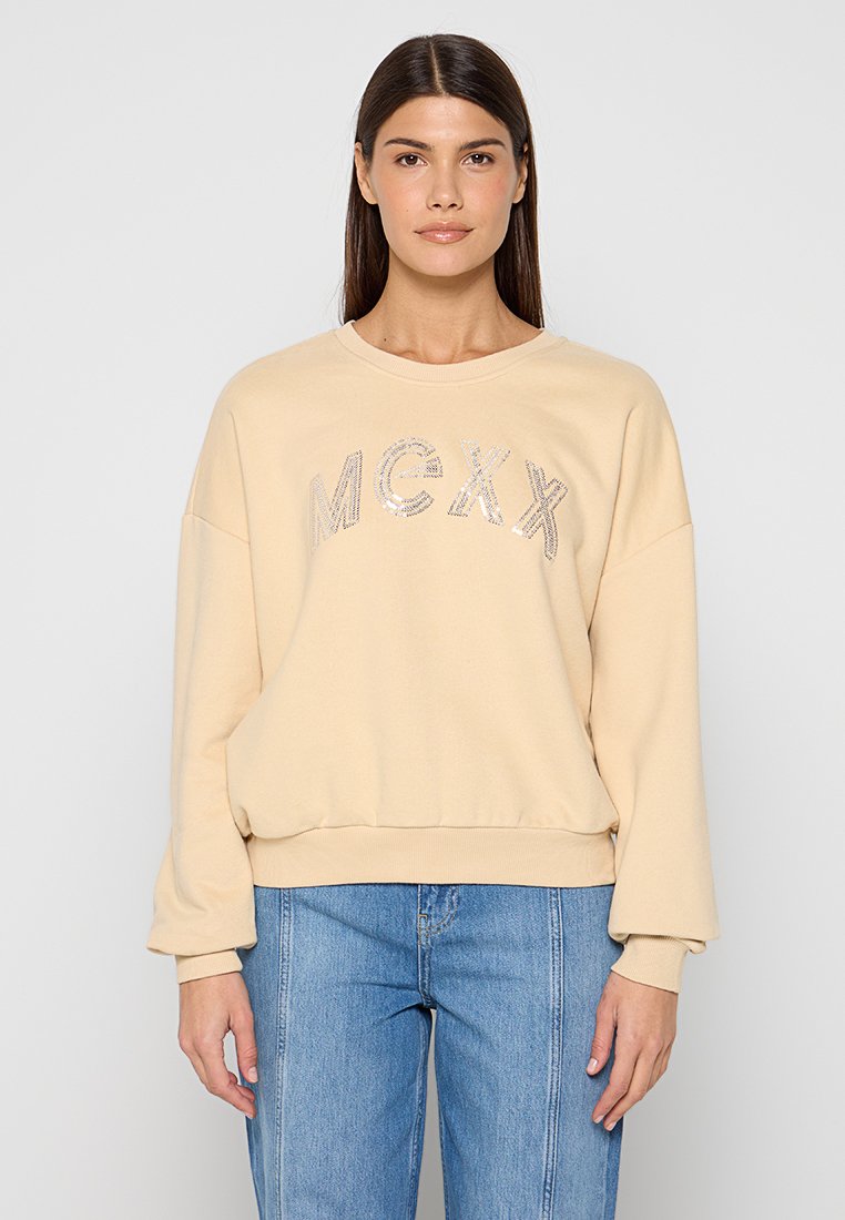 Beiger Pullover mit rundem Ausschnitt, überschnittenen Schultern und gerippten Bündchen. Auf der Vorderseite befindet sich eine metallische "MCXX"-Beschriftung. Kombiniert mit blauen Jeans.