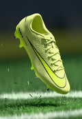 Helle gelbe Fußballschuhe mit strukturierten Oberflächen, schwarzen Akzenten und Schnürsenkeln. Über dem Gras schwebend, zeigen sie die geformten Stollen für Traktion.