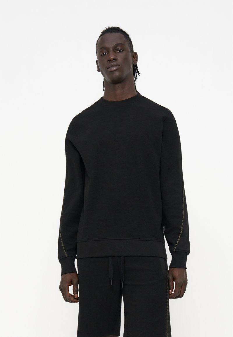 Große Person mit dunkler Haut und geflochtenem Haar, die einen schlichten schwarzen Langarm-Sweatshirt und schwarze Shorts trägt, steht vor einem weißen Hintergrund.