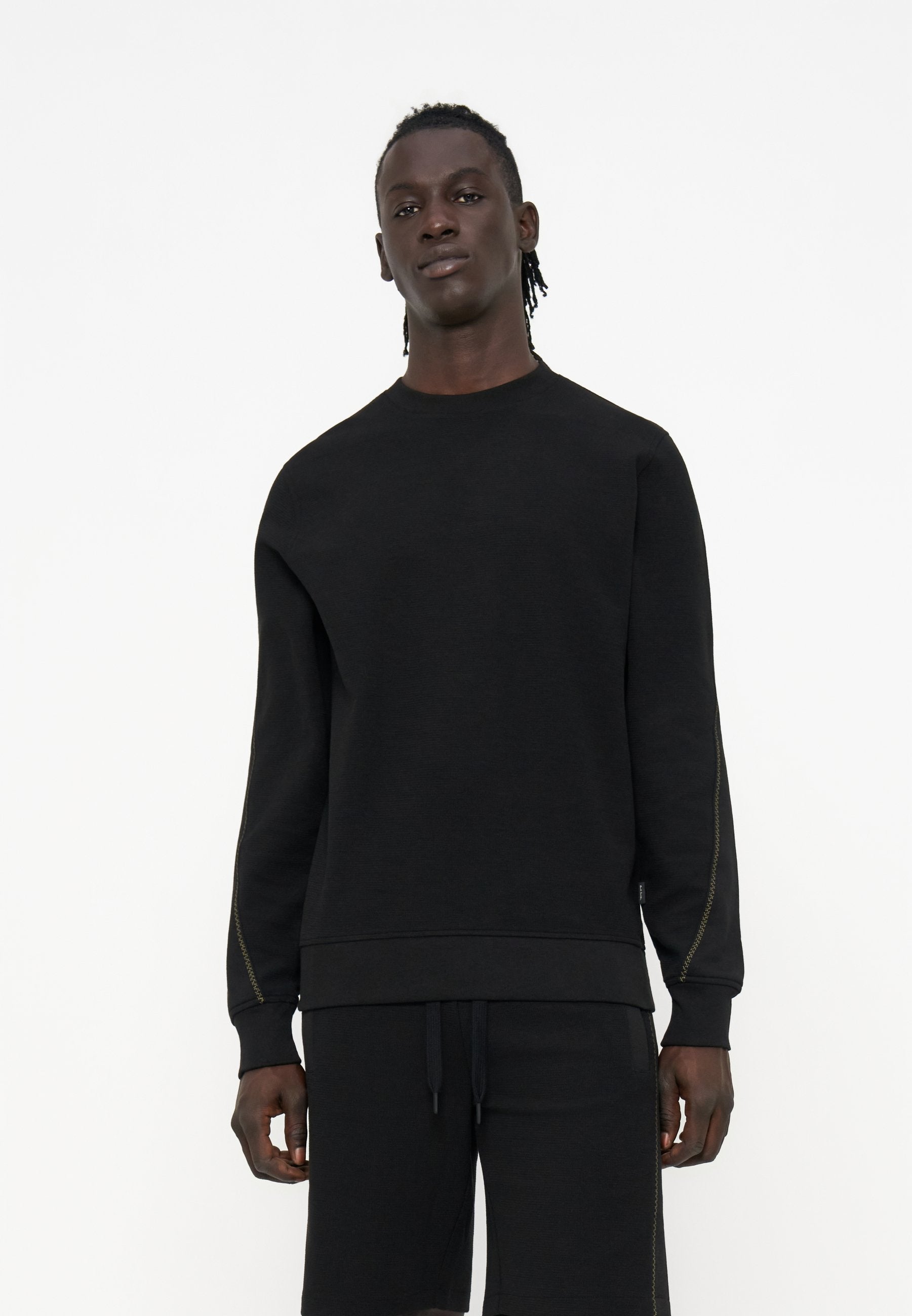 Große Person mit dunkler Haut und geflochtenem Haar, die einen schlichten schwarzen Langarm-Sweatshirt und schwarze Shorts trägt, steht vor einem weißen Hintergrund.