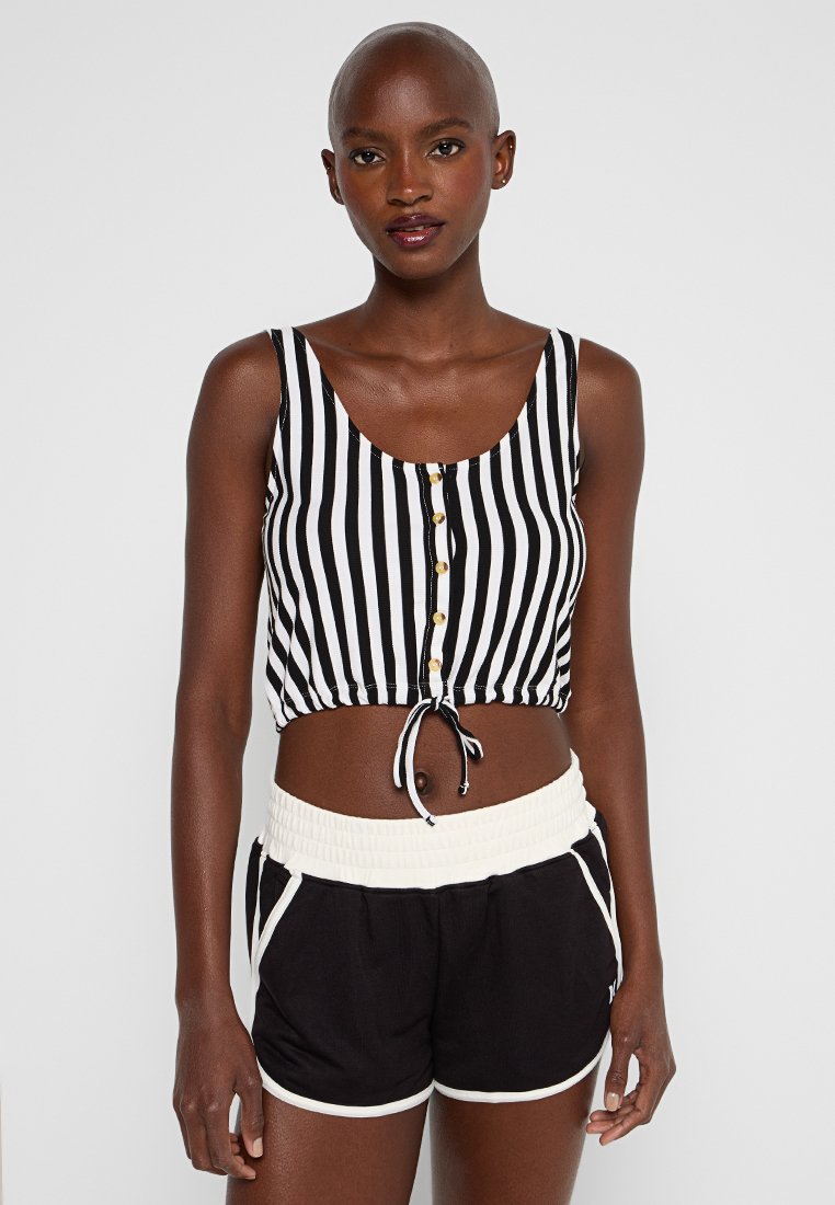 Frau in einem ärmellosen Crop-Top mit schwarzen und weißen vertikalen Streifen und Knöpfen sowie schwarzen Sportshorts mit weißem Saum.