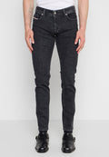 Mann trägt slim fit schwarze Jeans, schwarze Lederschuhe und ein weißes Hemd, steht vor einem schlichten hellen Hintergrund.