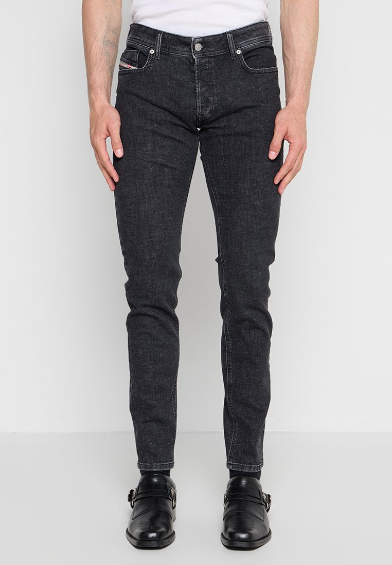 Mann trägt slim fit schwarze Jeans, schwarze Lederschuhe und ein weißes Hemd, steht vor einem schlichten hellen Hintergrund.