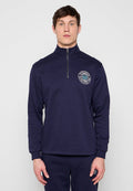 Mann trägt navyblauen Sweatshirt mit Viertel-Reißverschluss und passende Hose mit dem Logo "Worldwide Established 1969" auf Brust und Oberschenkel, steht vor weißem Hintergrund.