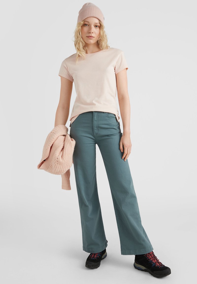 Helles pinkes Baumwoll-T-Shirt, türkisfarbene weit geschnittene Hose, ein beigefarbener Strickpullover über dem Arm und schwarze Wanderschuhe mit roten Akzenten.