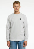 Hellgraues Sweatshirt aus strukturiertem Stoff, mit Rundhalsausschnitt und kleinem schwarzem Logo-Patch auf der Vorderseite. Kombiniert mit blauen Jeans.