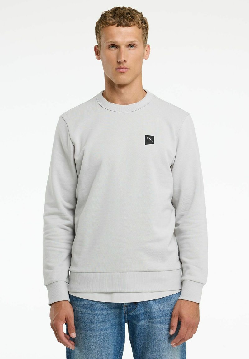 Hellgraues Sweatshirt aus strukturiertem Stoff, mit Rundhalsausschnitt und kleinem schwarzem Logo-Patch auf der Vorderseite. Kombiniert mit blauen Jeans.