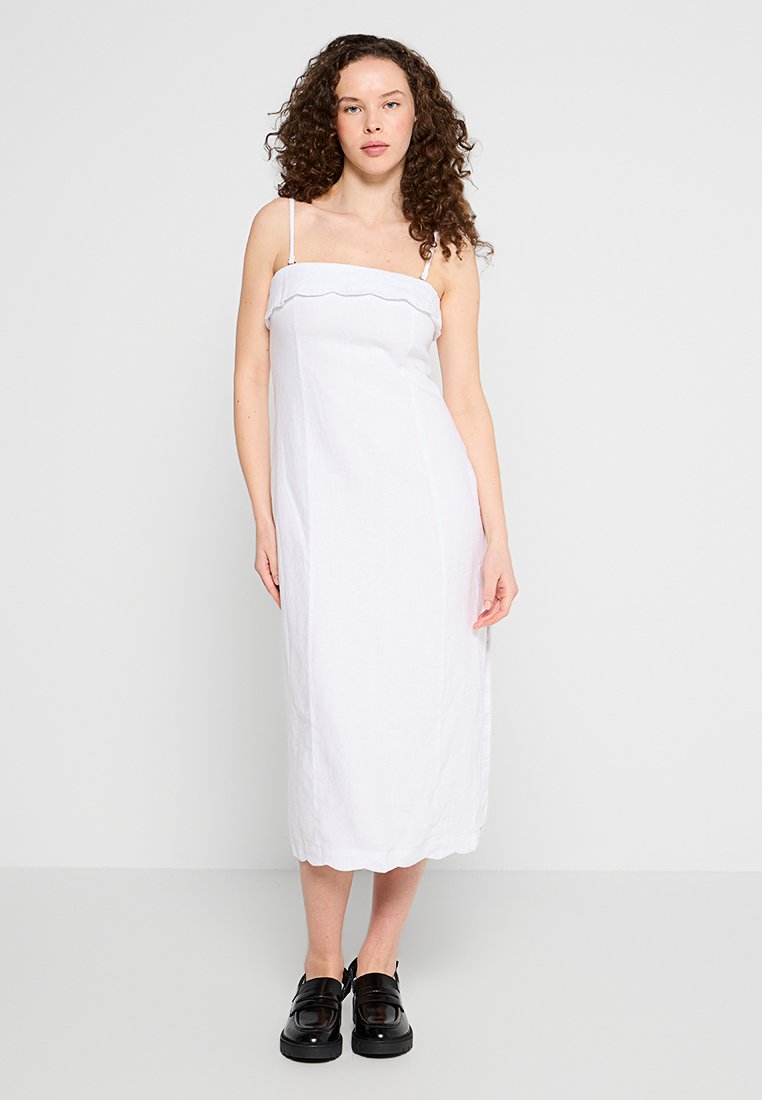 Weißes Leinen-Midikleid mit gewelltem Saum, dünnen verstellbaren Trägern und geradem Schnitt. Getragen mit schwarzen Plateauschuhen.