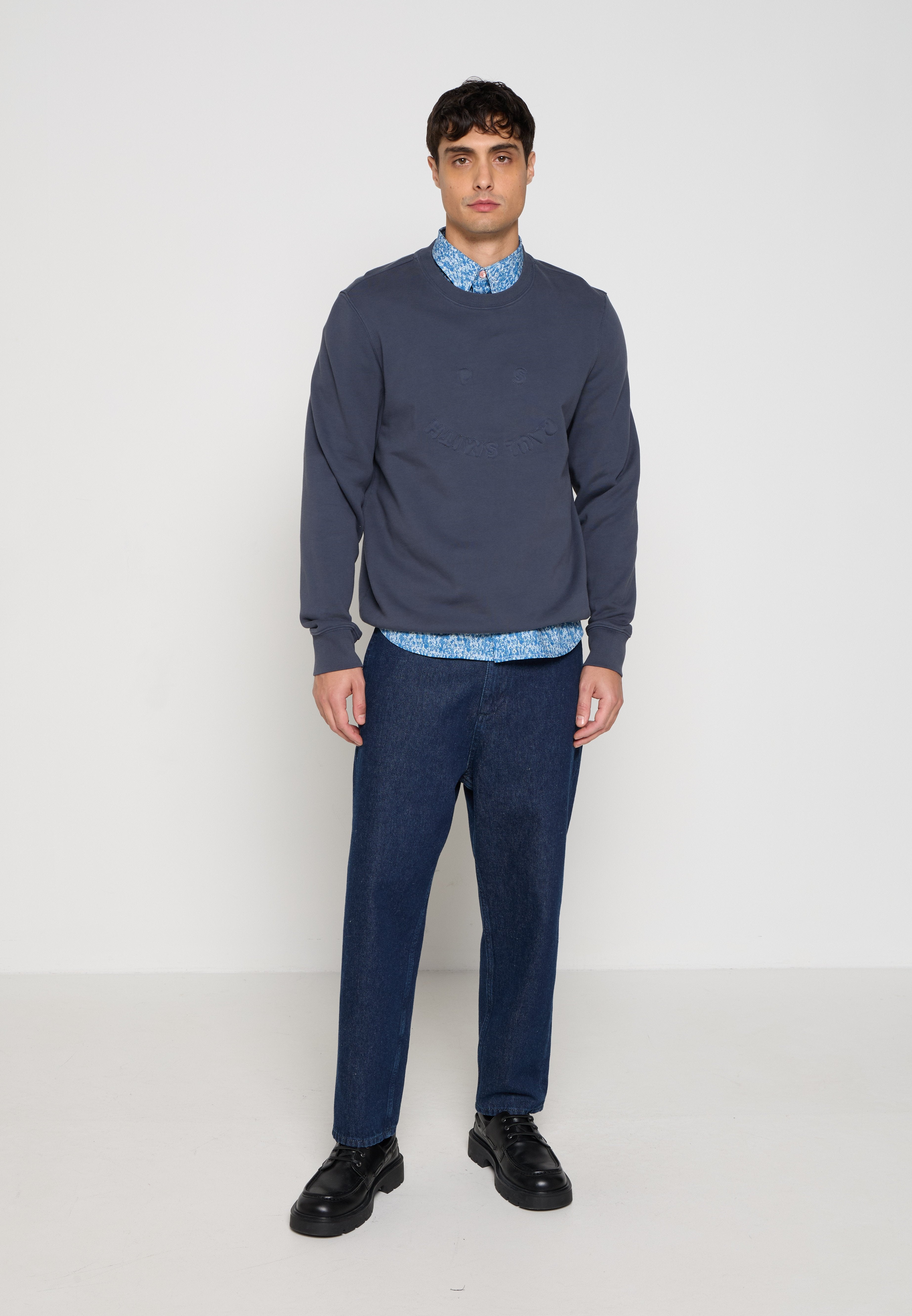 Mann trägt marineblauen Pullover über blau gemustertem Hemd, dunkelblaue Jeans und schwarze Lederschuhe, steht vor einem schlichten hellen Hintergrund.