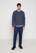 Mann trägt marineblauen Pullover über blau gemustertem Hemd, dunkelblaue Jeans und schwarze Lederschuhe, steht vor einem schlichten hellen Hintergrund.
