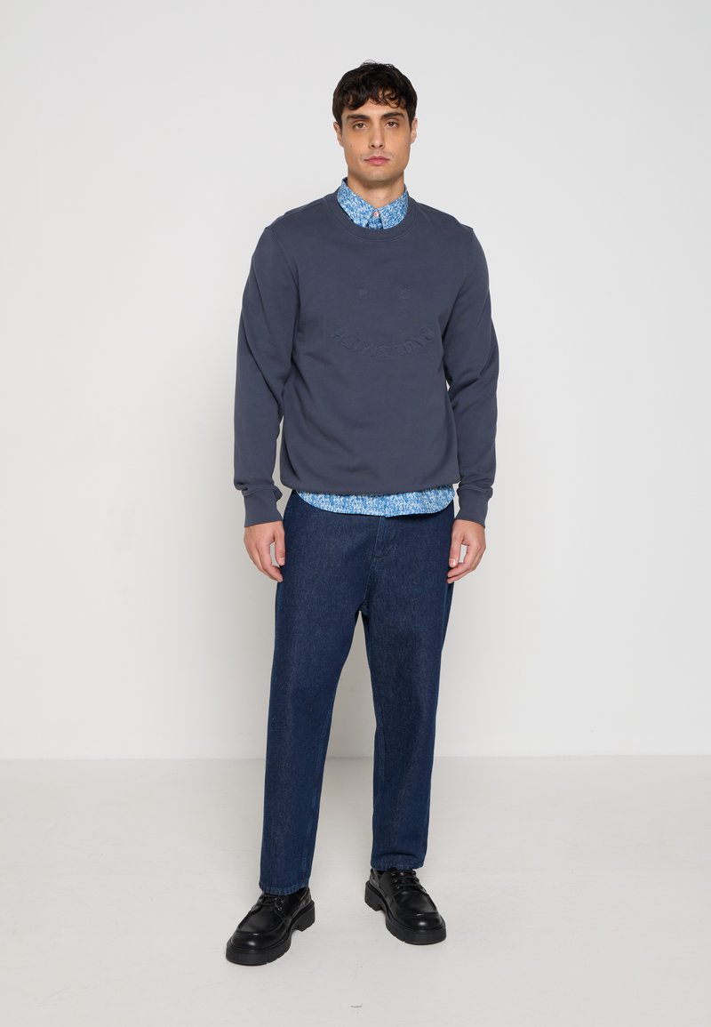 Mann trägt marineblauen Pullover über blau gemustertem Hemd, dunkelblaue Jeans und schwarze Lederschuhe, steht vor einem schlichten hellen Hintergrund.