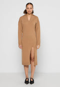 Strickkleid in camel mit V-Ausschnitt, langen Ärmeln, seitlichem Schlitz und geripptem Saum. Kombiniert mit schwarzen, spitz zulaufenden Schuhen.
