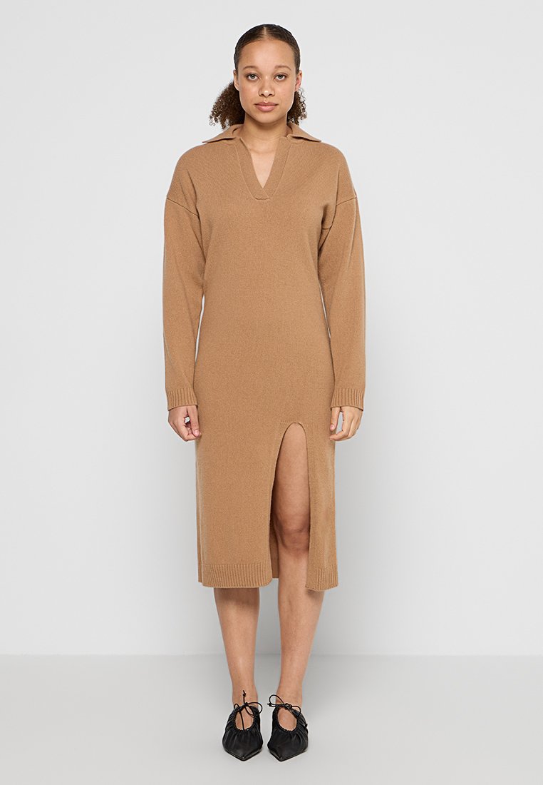 Strickkleid in camel mit V-Ausschnitt, langen Ärmeln, seitlichem Schlitz und geripptem Saum. Kombiniert mit schwarzen, spitz zulaufenden Schuhen.