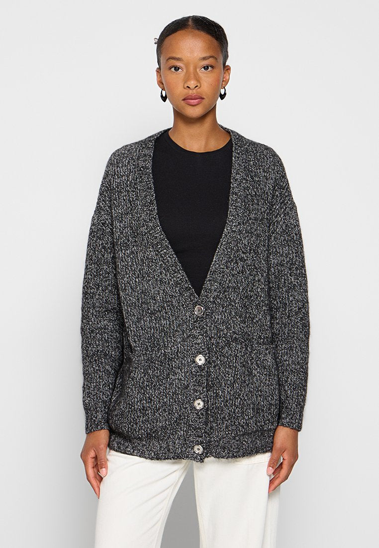 Frau in einem schwarzen Shirt, einer grauen, grob gestrickten, geknöpften Strickjacke, weißen Hosen und schwarzen Creolen, die vor einem schlichten Hintergrund steht.