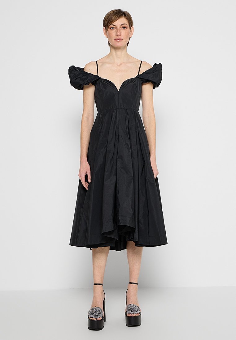 Frau trägt ein schwarzes, schulterfreies Midikleid mit Puffärmeln und schwarze Plateausandalen, die mit floralen Verzierungen geschmückt sind.