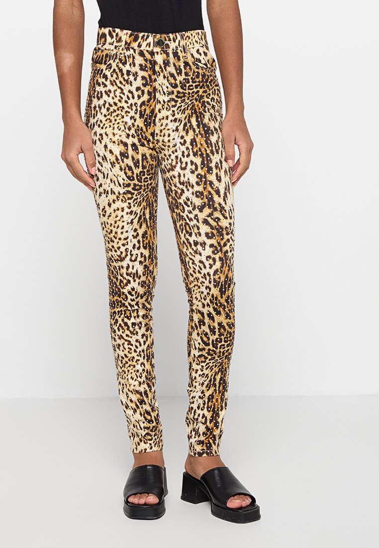 Schmal geschnittene Leopardenmuster-Hose mit hoher Taille und Strassakzenten, getragen mit schwarzen, offenen Sandaletten mit Absatz auf weißem Hintergrund.