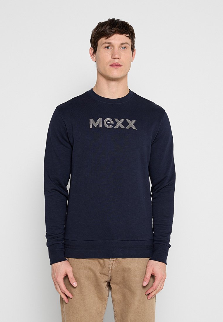 Mann trägt einen dunkelblauen Pullover mit langen Ärmeln und dem "MEXX"-Logo sowie eine hellbraune Hose, steht vor einem schlichten hellgrauen Hintergrund.